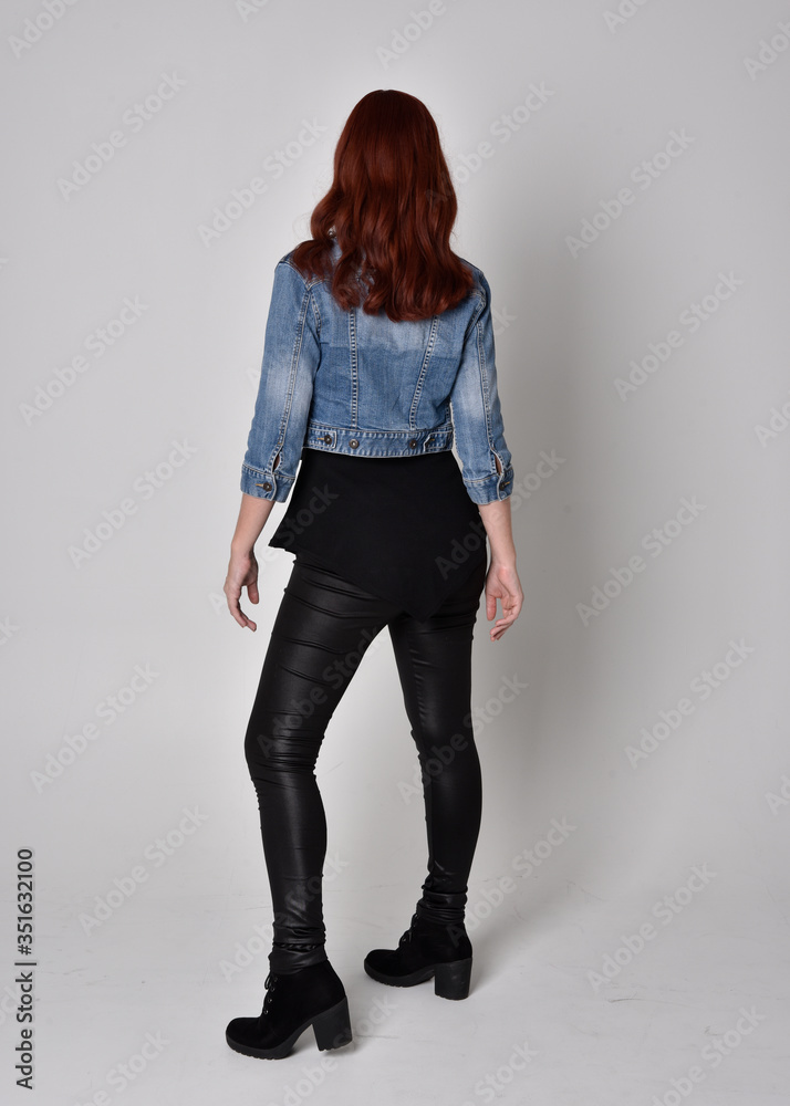 portrait of a pretty girl with red hair wearing black leather pants and denim jacket. Full length standing pose with back to the camera, isolated against a grey studio background portrait of a pretty girl with red hair wearing black leather pants and denim jacket. Full length standing pose with back to the camera, isolated against a grey studio background