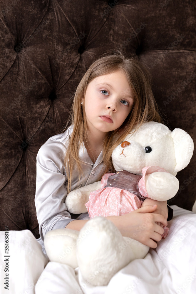 A little girl sadly hugs a toy in bed. The child is ill. A pretty girl with big eyes lies with a Teddy bear in a white bed and looks sadly at the camera.