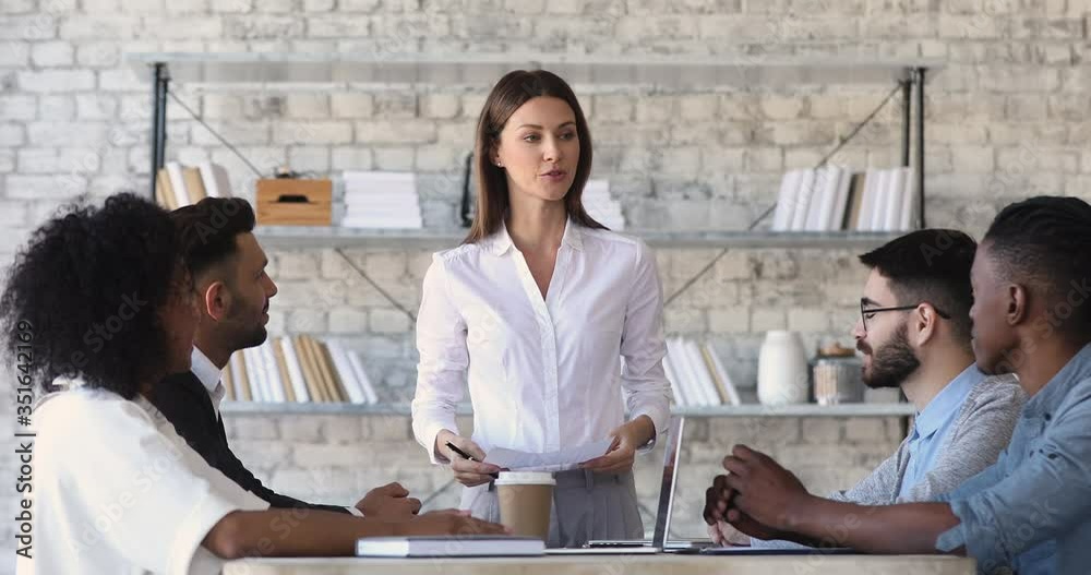 Confident female team leader boss supervisor standing at working desk, holding paper documents, talking to motivated interested focused young mixed race teammates colleagues in modern office.