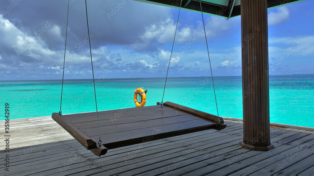 Obraz premium A delightful vacation in the middle of the ocean. A swing is suspended on a platform in the ocean. A place to relax in the shade and watch the colors of the aquamarine ocean and amazing cloudy scenery