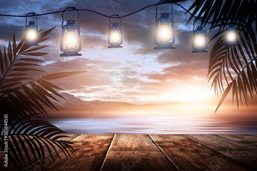 Garland of night lanterns on the pier, against the backdrop of a sea evening landscape with sunset. Palm tree branches, silhouettes, sunlight. Wooden table. Night view, open-air seascape.
