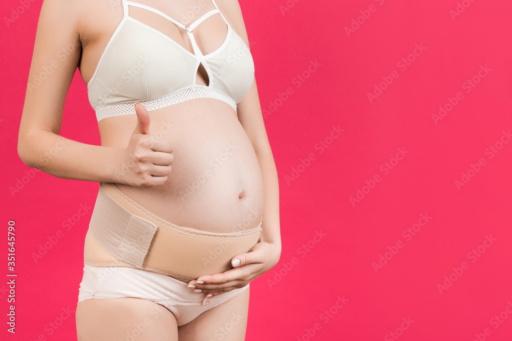 Close up of pregnant woman in underwear showing thumb up gesture and wearing maternity belt to reduce pain in the back at pink background with copy space. Orthopedic abdominal support belt concept Close up of pregnant woman in underwear showing thumb up gesture and wearing maternity belt to reduce pain in the back at pink background with copy space. Orthopedic abdominal support belt concept