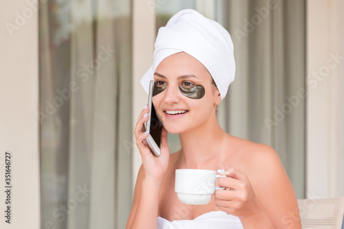 Beautiful woman wrapped in towel after shower with patches under eyes is drinking coffee and talking on the phone at the hotel terrace