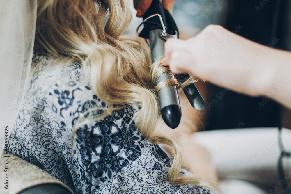 Fototapeta premium Hair curler. Hair stylist curling blonde hair with professional equipment closeup, morning preparations for wedding day. Bride in hair salon styling her hair