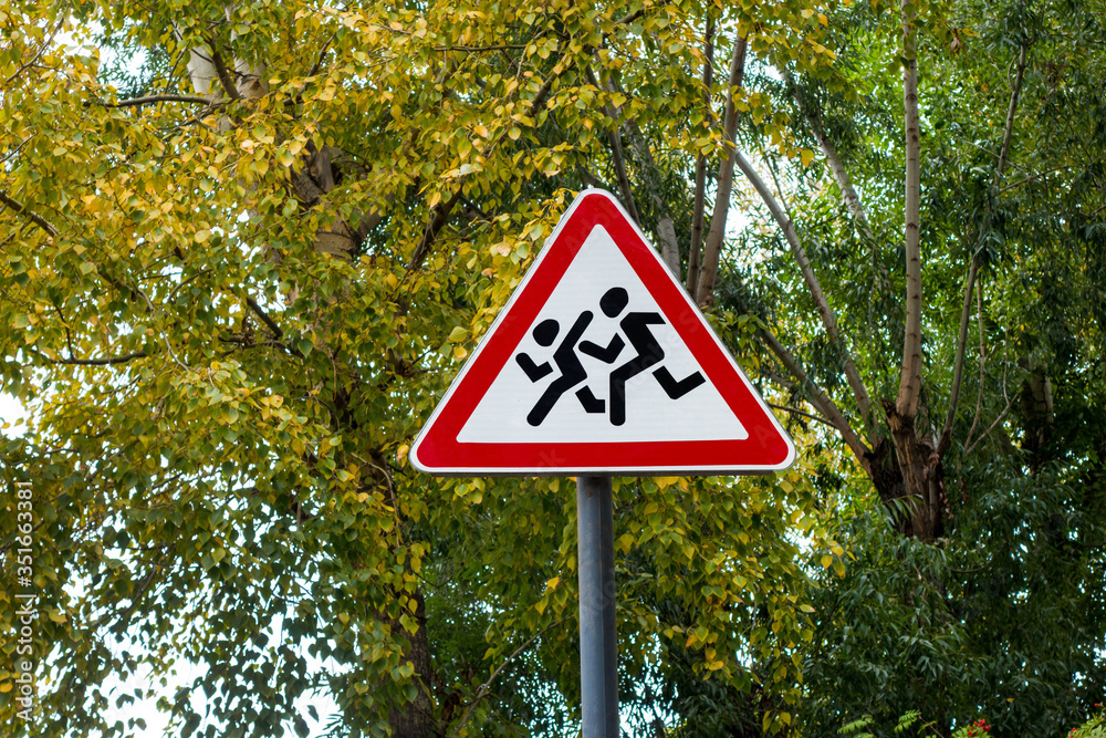International traffic sign 'Children crossing', standing near urban park zone. Trees are on background