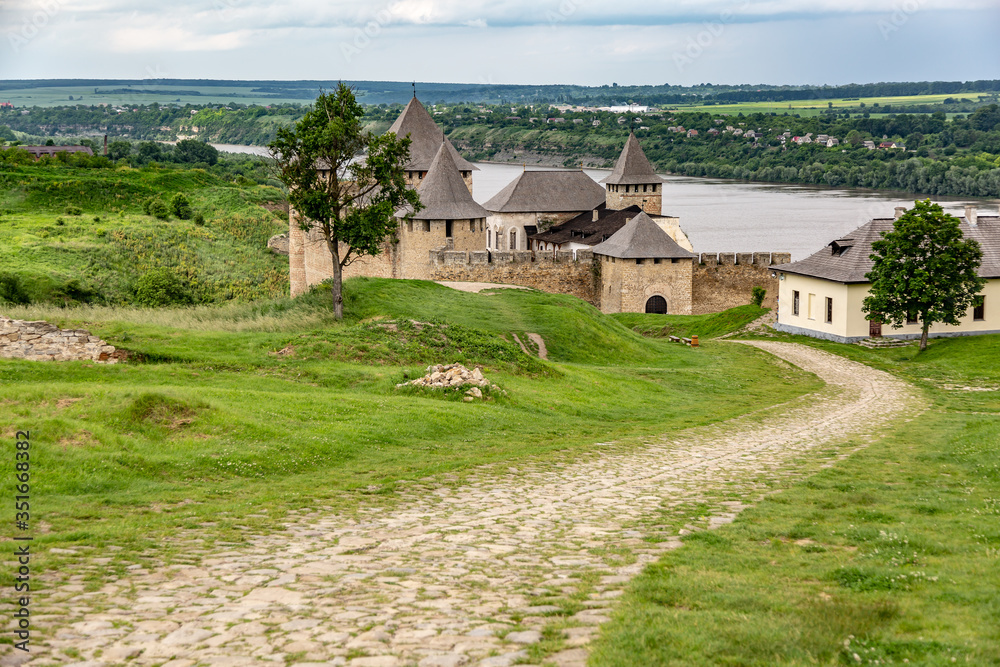 Khotyn Fortress castle in Ukraine and the road to it, river on a background of dark clouds on a cloudy windy day in summer. Horizontal orientation.
