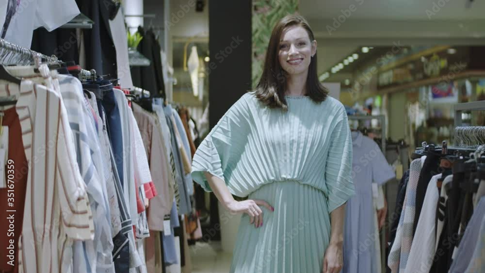 Cheerful brunette lady in dress is standing with hands on her hip and looking at the camera in clothes shop. Front view of smiling stylish woman posing for the camera in the shop. Cheerful brunette lady in dress is standing with hands on her hip and looking at the camera in clothes shop. Front view of smiling stylish woman posing for the camera in the shop.
