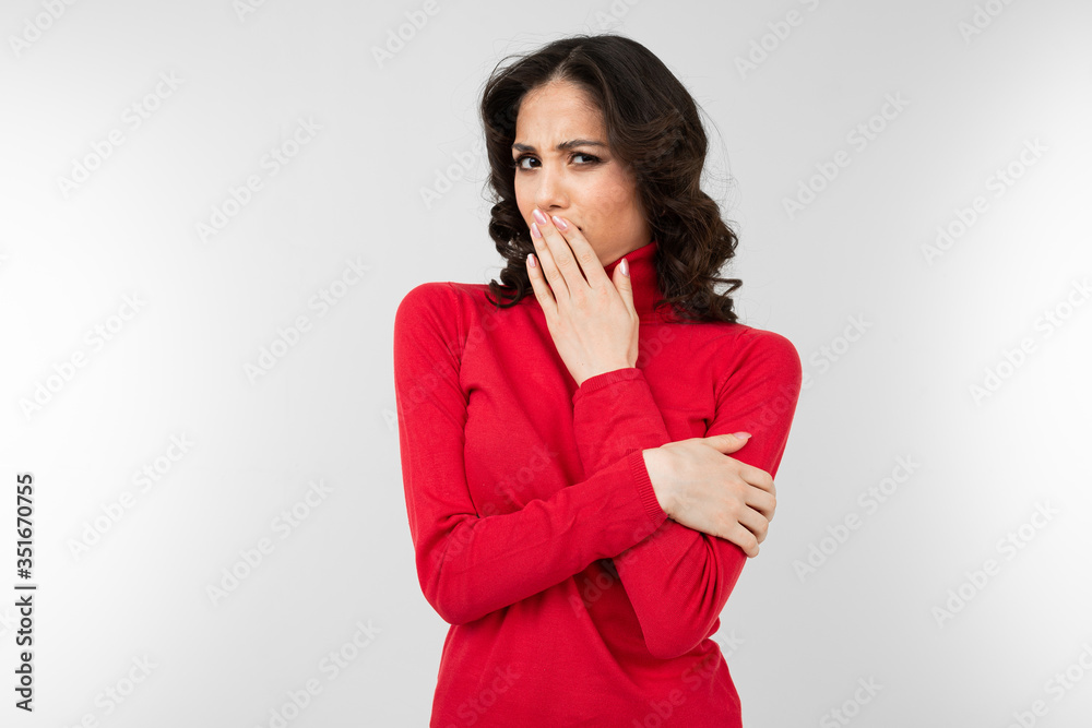 brunette girl in a red tight-fitting sweater hides a secret by closing her mouth with her hand on a white background with copy space brunette girl in a red tight-fitting sweater hides a secret by closing her mouth with her hand on a white background with copy space