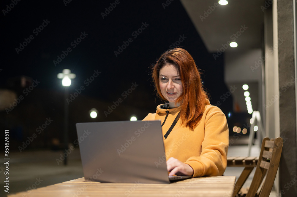 A woman works remotely at a laptop in a summer cafe late in the evening. Happy Girl studying while sitting on an empty street at a wooden table. Female freelancer in a sweatshirt at the evening A woman works remotely at a laptop in a summer cafe late in the evening. Happy Girl studying while sitting on an empty street at a wooden table. Female freelancer in a sweatshirt at the evening