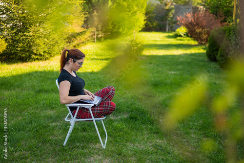 A woman with glasses sits in a folding chair with her legs crossed in a lotus position and typing on a laptop. Female freelancer remotely works in the garden of the cottage during quarantine.