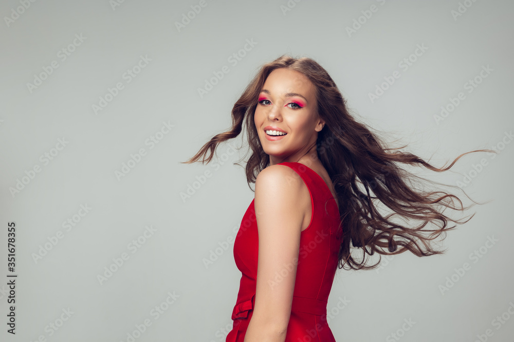 Fototapeta premium Beautiful young woman with long healthy curly hair and bright make up wearing red dress isolated on grey studio backgroud. Beauty and fashion, hair and skin care, treatment concept. Well-kept girl.