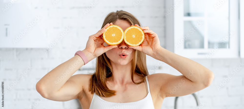 Young charming woman holding two halves of oranges near her beautiful face in the kitchen background. Healthy food concept. Young charming woman holding two halves of oranges near her beautiful face in the kitchen background. Healthy food concept.