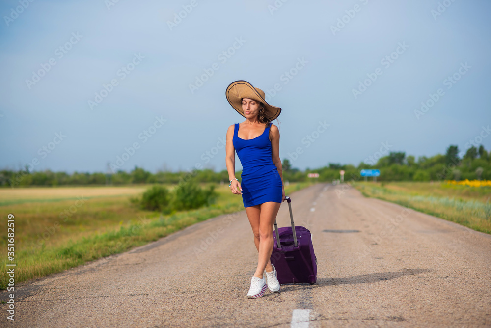 A woman in a hat and with a big bag is walking along the roadway. Girl in a blue tight-fitting dress with a purple suitcase on the track. Brunette with a sports figure on the road.