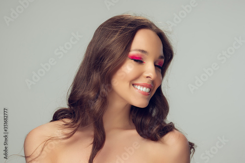 Close up of beautiful young woman with long healthy curly hair and bright make up isolated on grey studio backgroud. Beauty and fashion, hair and skin care, treatment concept. Well-kept healthful girl