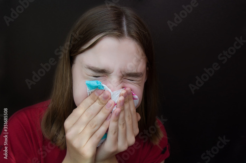 Sick young girl with allergies blows her nose in a napkin. Dark background. Copyspace.