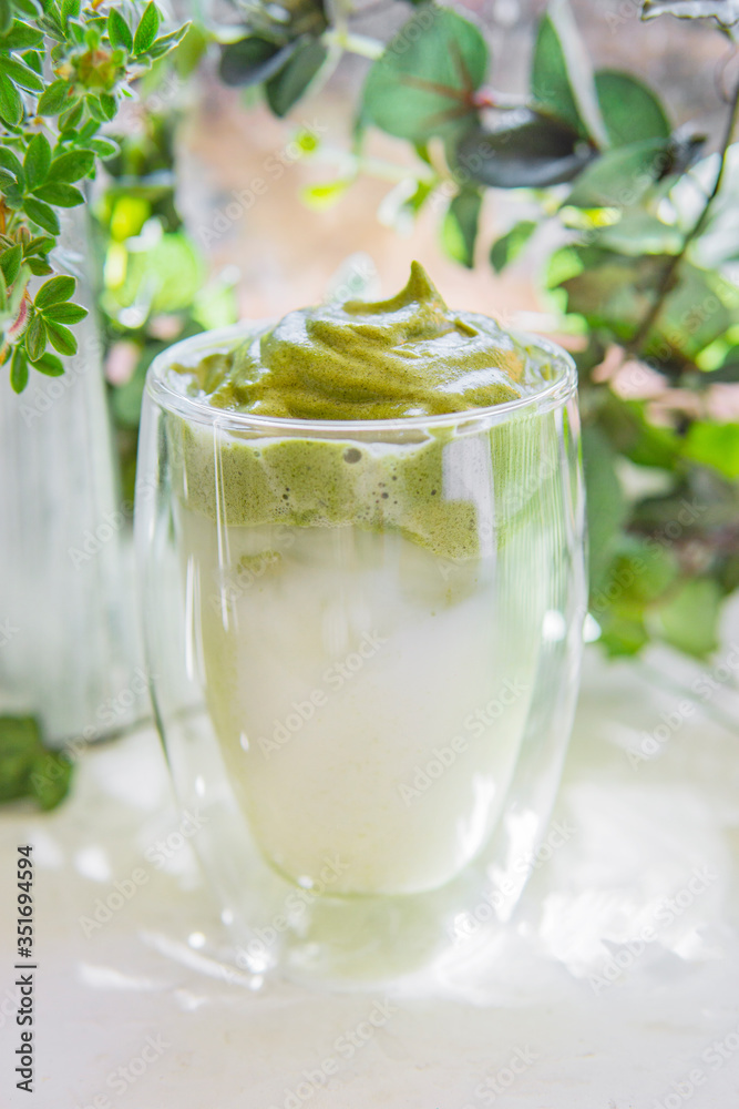 Dalgona matcha coffee or tea - green iced coffee drink with milk on white table with plants, sunny light. New trend drink in social media. Morning healthy reverse latte. Selective focus, copy space. Dalgona matcha coffee or tea - green iced coffee drink with milk on white table with plants, sunny light. New trend drink in social media. Morning healthy reverse latte. Selective focus, copy space.