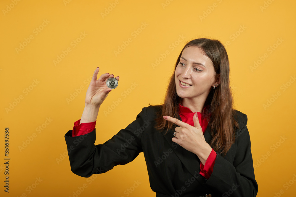 Young Attractive Woman Brunette In Black Stylish Suit, Red Shirt On Yellow Background, Successful Woman Holds A Bitcoin Coin In Her Hand, Smiles. The Concept Of Wealth, Business And E-Commerce Young Attractive Woman Brunette In Black Stylish Suit, Red Shirt On Yellow Background, Successful Woman Holds A Bitcoin Coin In Her Hand, Smiles. The Concept Of Wealth, Business And E-Commerce