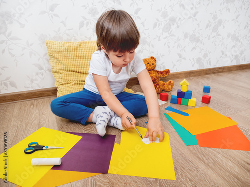 Toddler boy learns to cut colored paper with scissors. Kid sits on floor in kids room with toy blocks. Educational classes for children. Developing feeling sensations and fine motor skills at home. photo