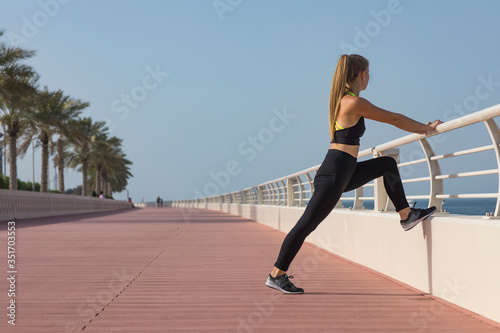 Portrait of fit and sporty young woman doing stretching in city. Young fitness woman runner stretching legs before run on city. Fitness runner doing warm-up routine on beach before running. Dubai