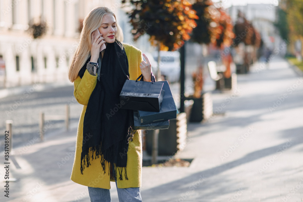 cute attractive blonde woman with packages on the street in sunny weather. communicates on the phone after shopping, positive emotions.