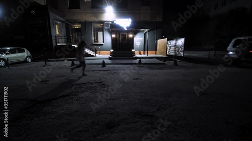 Behind the SUV, a woman in a medical coat is running. In the background is a bar building. Night. Overall plan.