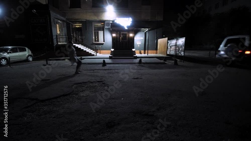 Behind the SUV, a woman in a medical coat is running. In the background is a bar building. Night. Overall plan.