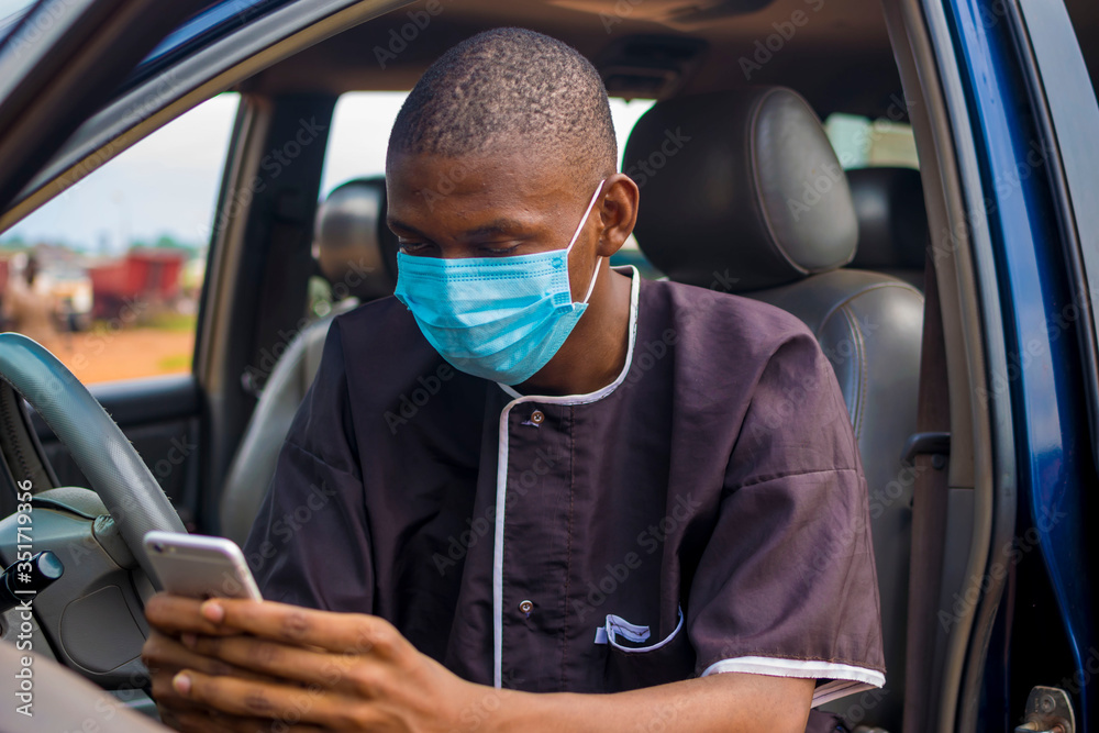young black man wearing a nose mask, sitting in his car and using his smartphone with his car door open