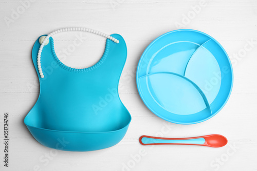 Colorful plastic dishware and silicone bib on white wooden table, flat lay. Serving baby food
