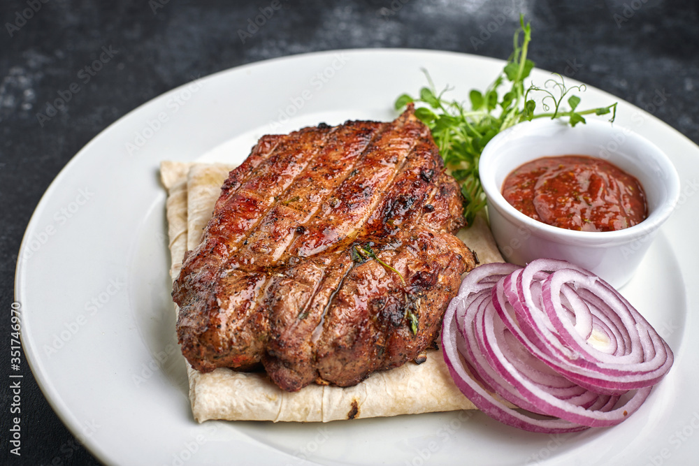 Grilled meat steak with onions, sauce and microgreen, on pita bread, on a white plate, against a dark background. space for text.