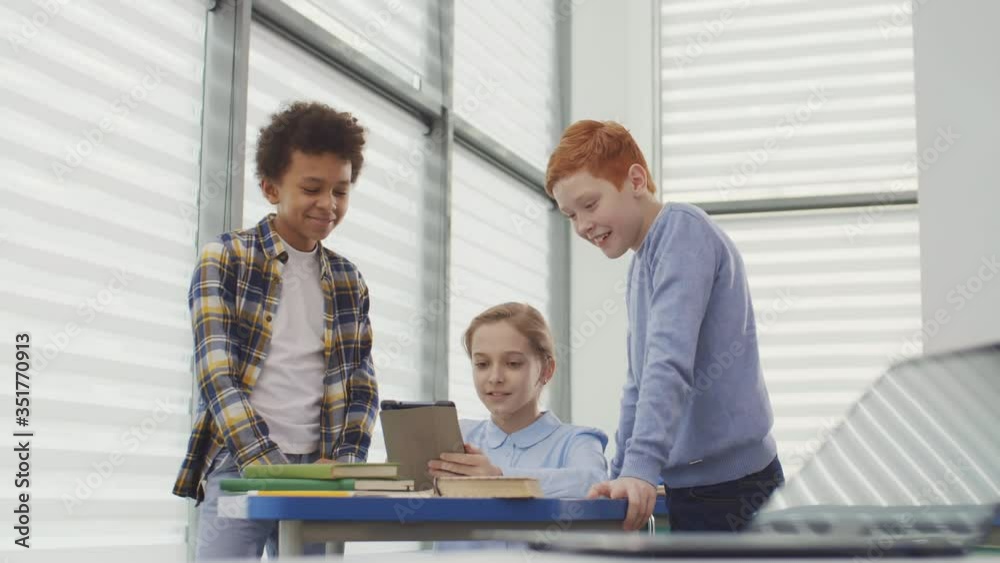 Lockdown of Caucasian girl sitting at desk in classroom, using tablet and talking to two diverse boys standing nearby