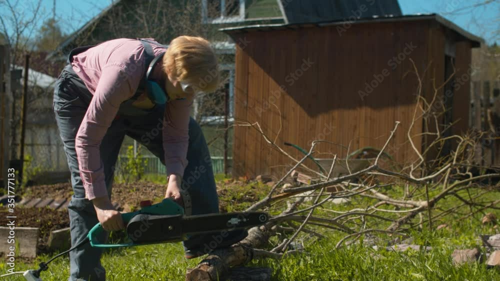 Mature woman cutting tree with an electric chain saw on the courtyard of a country house. Active and healthy lifestyle concept Mature woman cutting tree with an electric chain saw on the courtyard of a country house. Active and healthy lifestyle concept