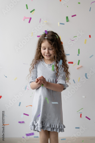 Charming curly brunette girl in a beautiful white dress with her hair is having fun, clapping her hands, playing with multi-colored falling confetti. Happy childhood. A little party .