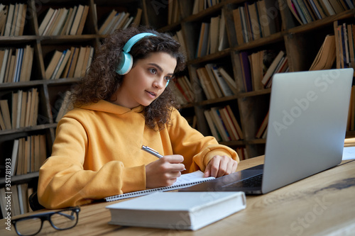 Hispanic teen girl, latin young woman school college student wear headphones learn watching online webinar webcast class looking at laptop elearning making notes or video calling remote teacher. photo