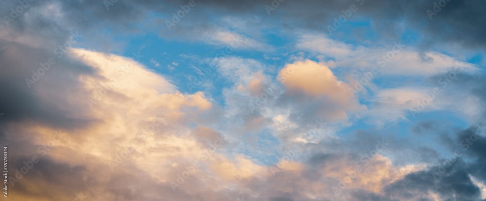 Beautiful colorful panoramic view of summer golden fluffy clouds illuminated by disappearing rays at sunset, dramatic dark thunderclouds in blue sky. Dramatic panorama of natural weather background