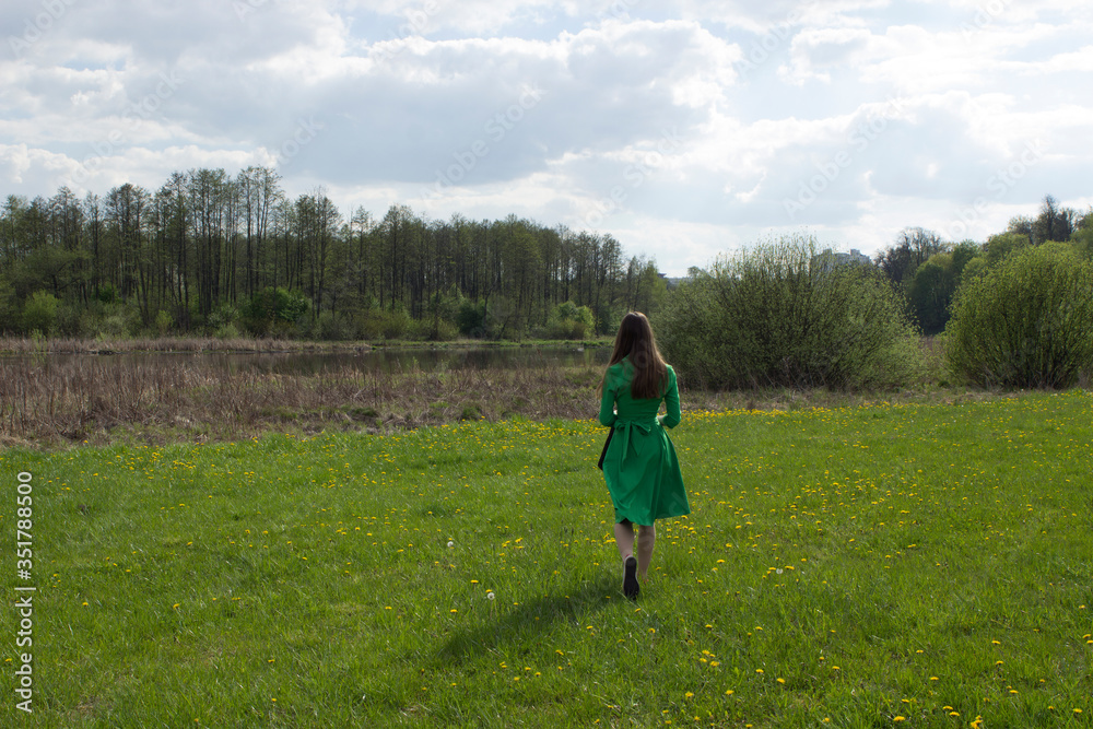 A lonely girl with long hair in a green dress goes to the river in the forest, meadow. Self-isolated girl in the park.