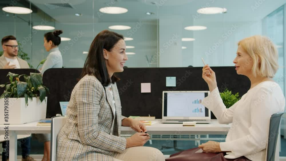 Cheerful women coworkers talking discussing work looking at laptop screen sitting at desk in open space office room. People and communication concept. Cheerful women coworkers talking discussing work looking at laptop screen sitting at desk in open space office room. People and communication concept.
