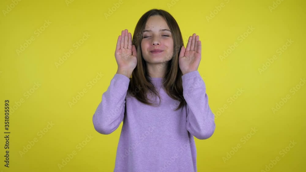 Teen girl in a pink sweater picks up palms from her face and smiles on a yellow background with copy space. Place for text or product. 4K slow motion footage Teen girl in a pink sweater picks up palms from her face and smiles on a yellow background with copy space. Place for text or product. 4K slow motion footage