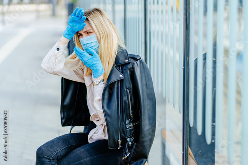 Unhealthy blonde fashionable woman traveler millennial on bus or tram stop wearing protective mask and gloves, ill, sick, from virus and air pollution