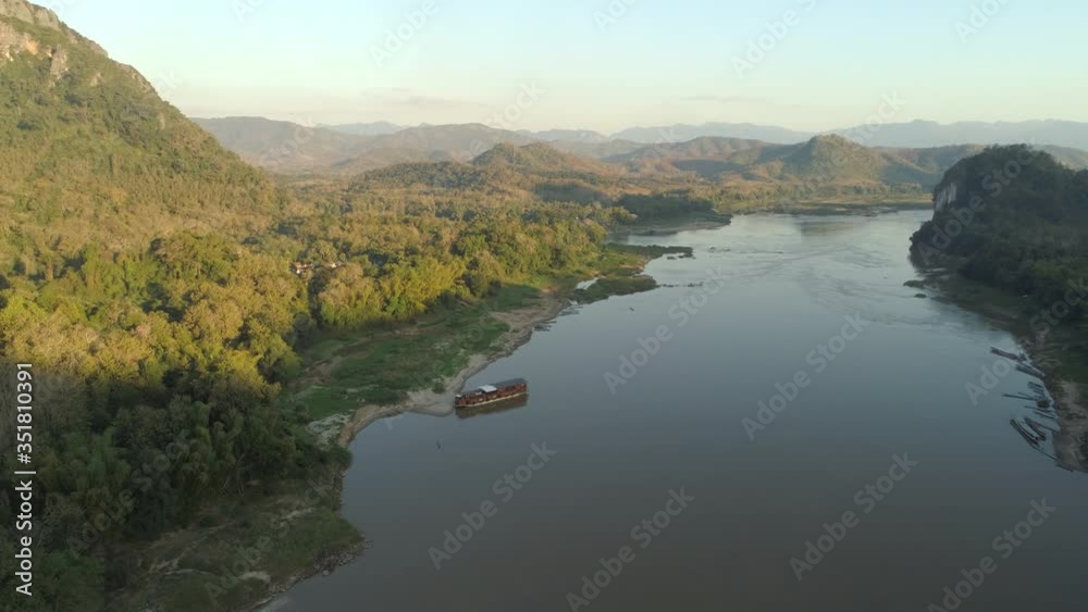 Aerial shot of cruise ship at riverbank against sky on sunny day, drone flying forward over river near mountains - Mekong River, Laos Aerial shot of cruise ship at riverbank against sky on sunny day, drone flying forward over river near mountains - Mekong River, Laos