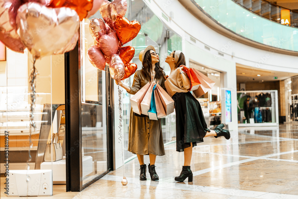 Obraz premium Shopping and entertainment, mall inside. Two beautiful girls with paper bags in the mall, heart-shaped balloons. The joy of consumption, Gift shopping, holiday.