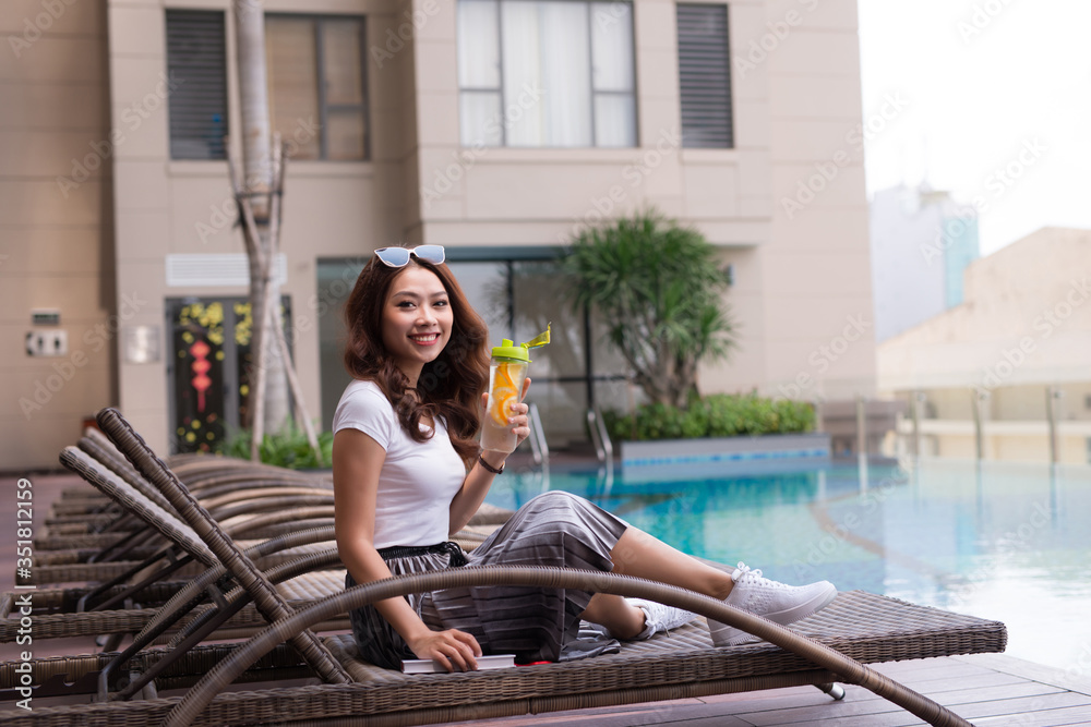 Young Asian woman sitting on deckchair and drinking juice near swimming pool