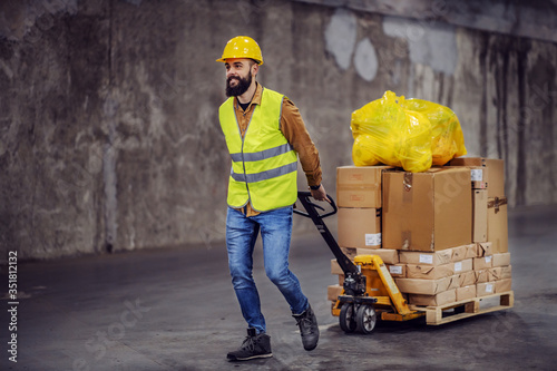 Full length of smiling hardworking bearded worker in vest, with safety helmet on head pulling a pallet truck with boxes, sack and building material. Building in construction process interior. photo