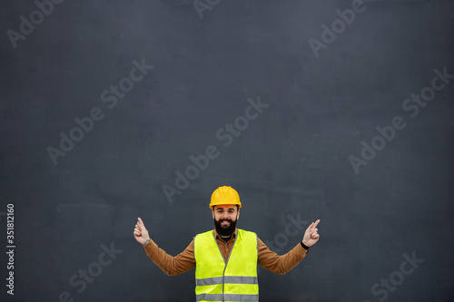 Young smiling attractive bearded worker in vest, with safety helmet on head standing in front of gray wall, looking at camera and pointing up with both hands.