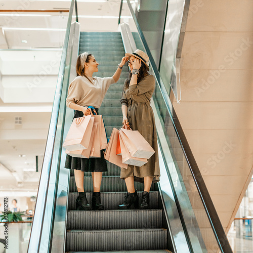 Two girls on an escalator in a shopping center. In the hands of paper bags. Shopping, leisure and entertainment.