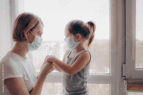 Family Mom and Daughter in Medical Mask. Young Woman and Child little girl sitting by the Window in Protective Masks against the Virus. Mom carefully corrects the mask on the face of the child. 