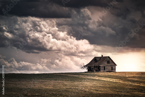 Old wooden farmhouse in the countryside at sunset with storm clouds in the sky. There is a short grass meadow around the house.