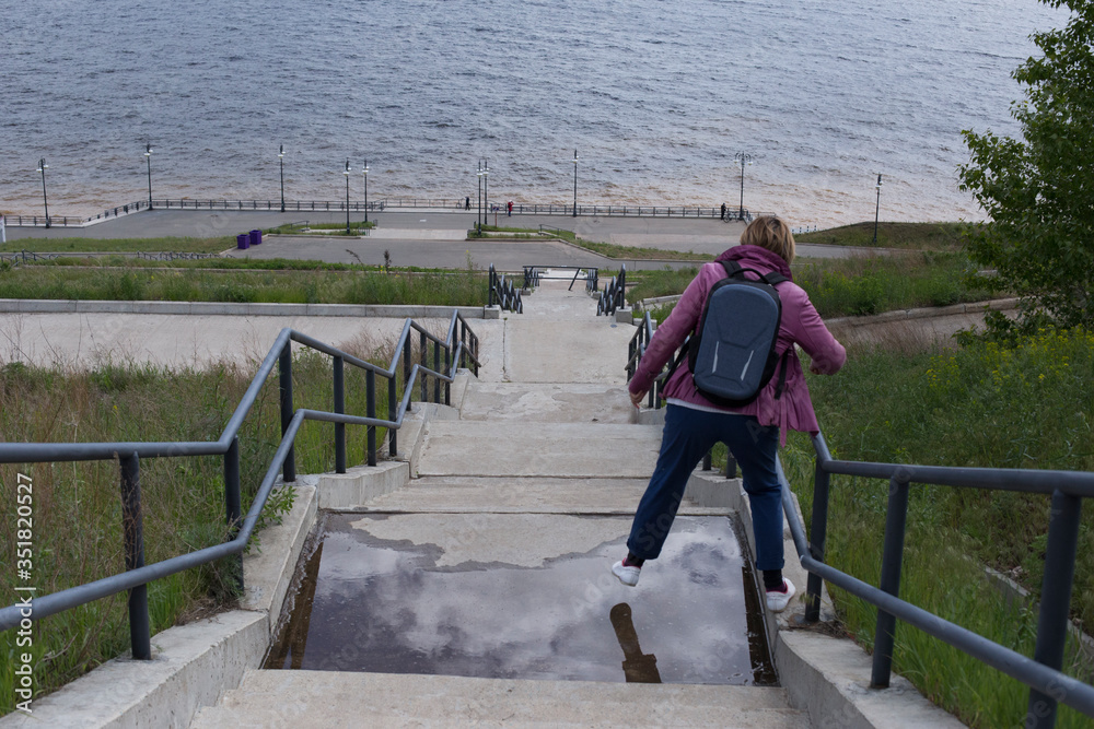 River embankment and high staircase. A woman is jumping through a puddle. A shot from the back high above the shore.
