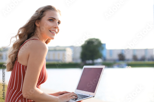 Portrait of pretty young woman sitting on riverbank during summer day, using laptop