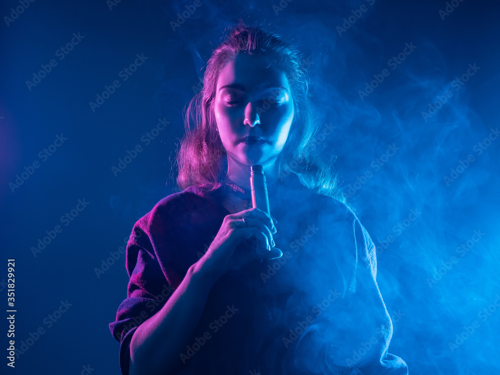 Obraz premium VAPE Smoking. A girl with closed eyes smokes a VAPE. The pleasure of Smoking an electronic cigarette. VAPE shop. Gadgets for Smoking. The concept of Smoking an e-cigarette in blue.