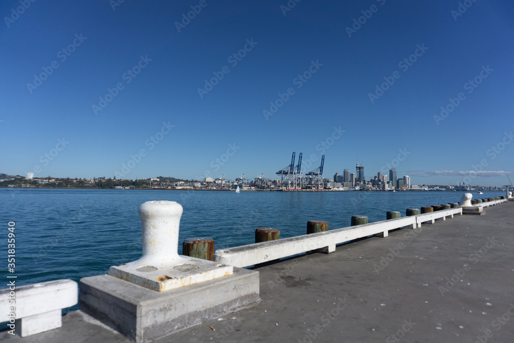 Auckland skyline and port with container cranes on other side of harbour with Devonport wharf and white bollard in foreground.
